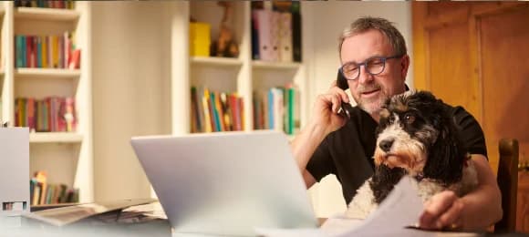 A freelancer on a phone call at his desk at home, with his dog beside him